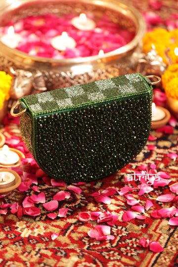 A green beaded handbag with a silver and green checkered pattern on the flap, styled on a patterned rug with pink flower petals, candles, and a brass pot in the background.