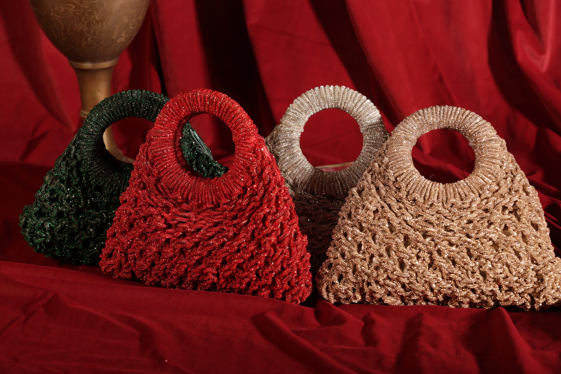 Four shimmering mesh-woven Glimora bags in jewel and metallic tones arranged against rich red fabric.