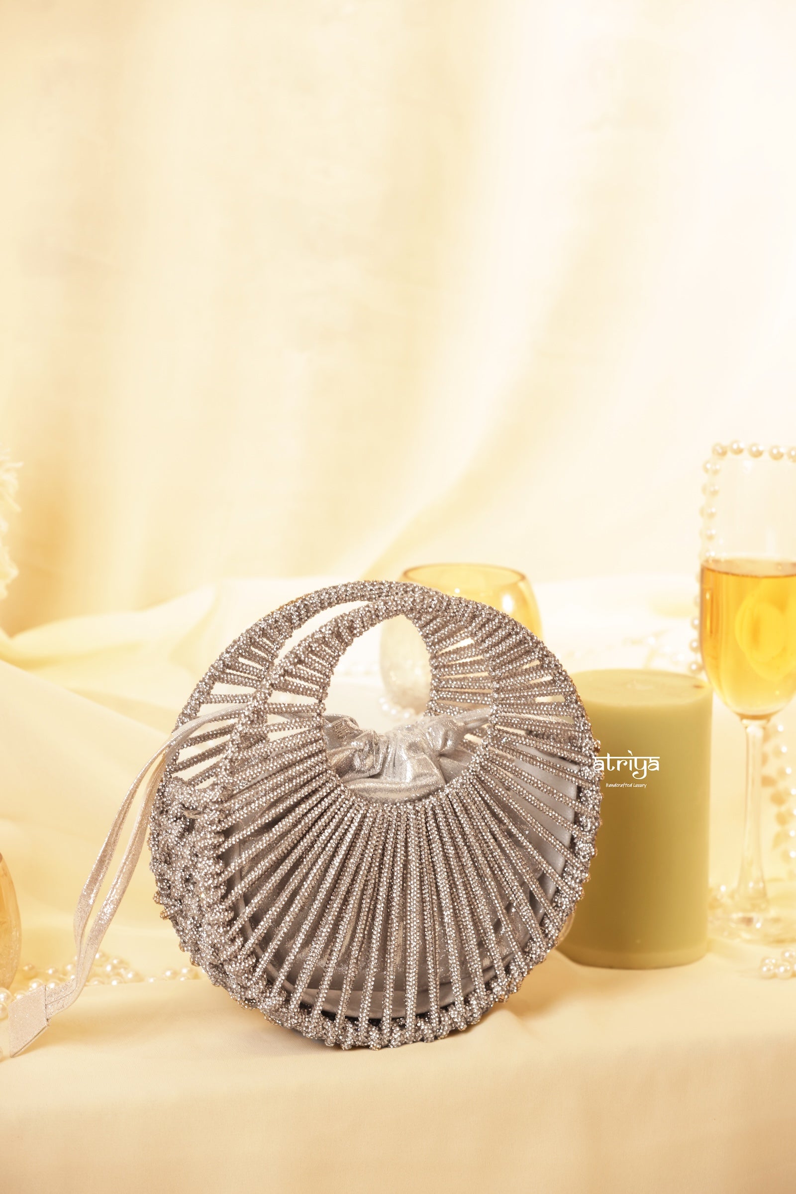 A round silver beaded evening clutch bag with a top handle, styled on a cream draped tablecloth with candles and glasses in the background.