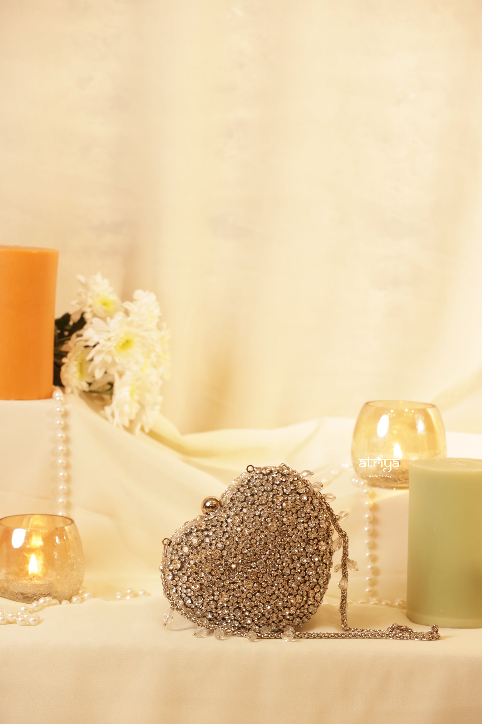 A side profile shot of the closed Heartbreaker gold stone clutch by Atriya Jaipur. The heart-shaped silhouette is fully covered in reflective gold stones with a secure metallic clasp at the top and an elegant, detachable gold chain resting on a luxurious white silk setting.
