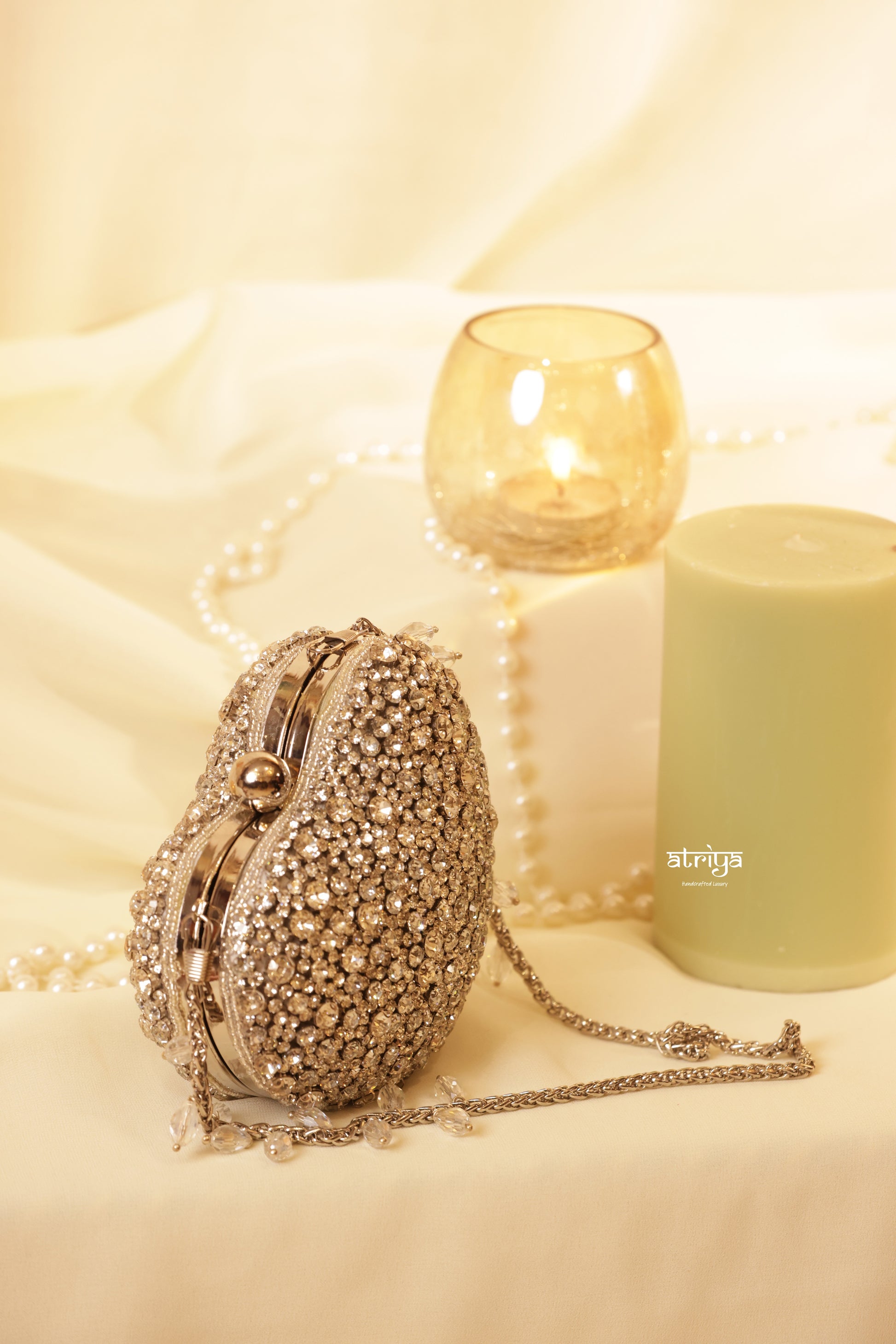 An open gold heart-shaped evening clutch bag by Atriya Jaipur, heavily encrusted with shimmering gold stones on the exterior. The bag reveals a smooth tan suede interior lining and a luxury gold chain strap with stone droplets, styled on a white fabric background next to a lit candle and pearl strands.