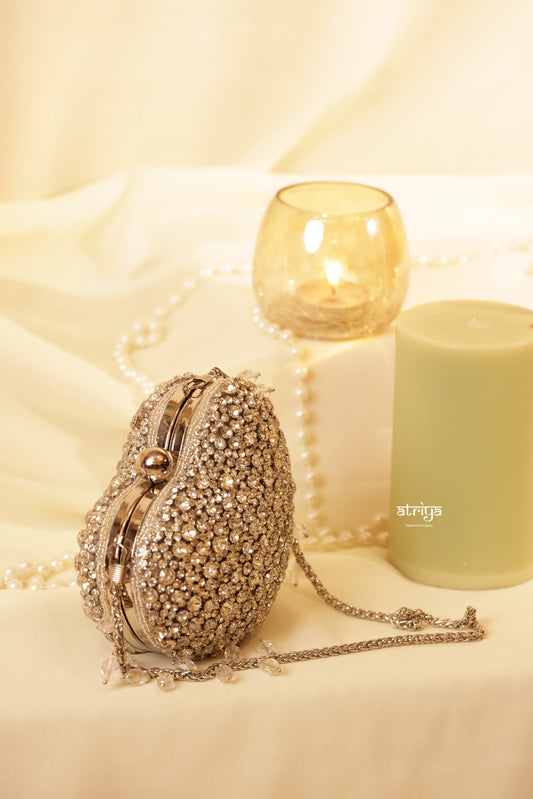 An open gold heart-shaped evening clutch bag by Atriya Jaipur, heavily encrusted with shimmering gold stones on the exterior. The bag reveals a smooth tan suede interior lining and a luxury gold chain strap with stone droplets, styled on a white fabric background next to a lit candle and pearl strands.