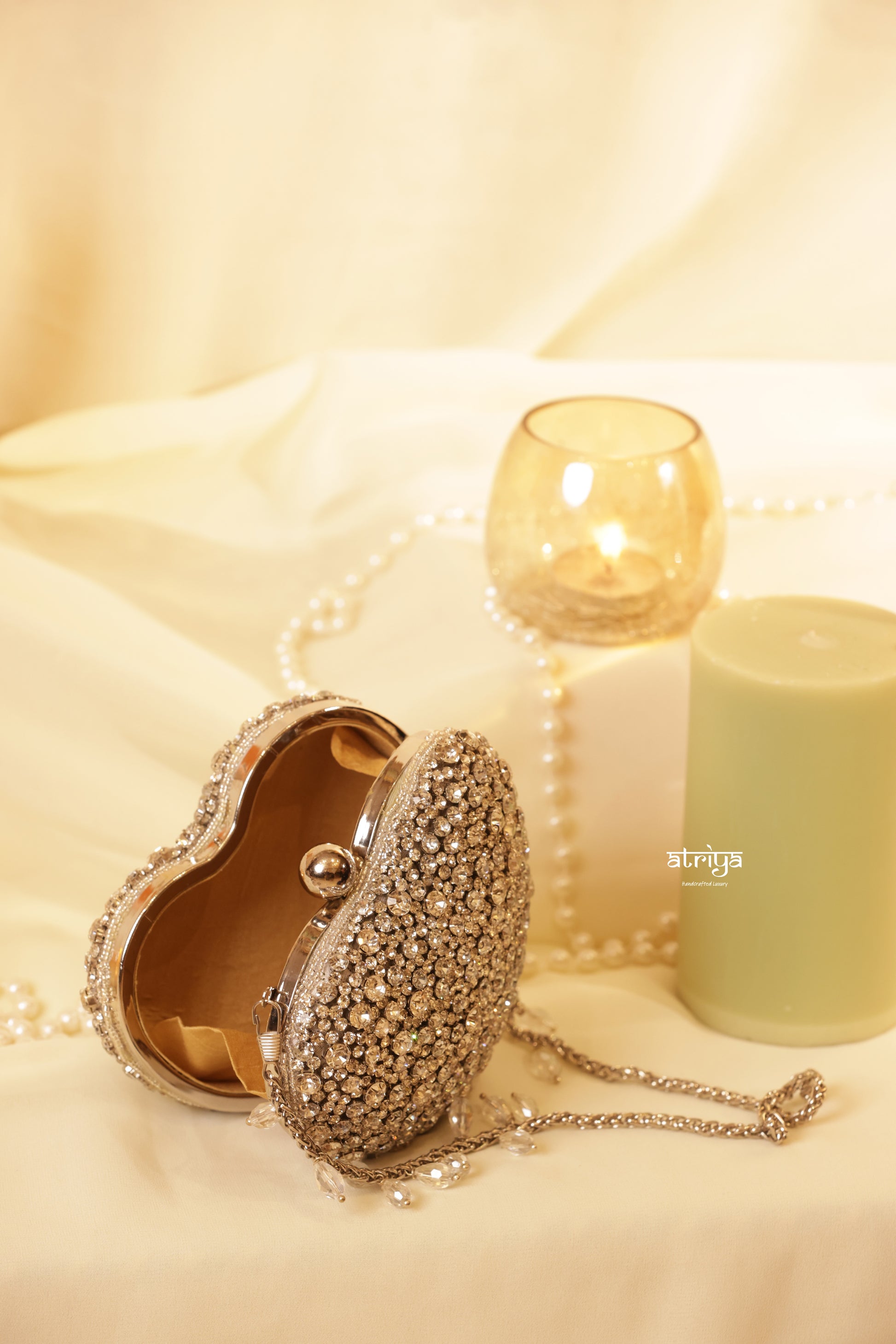 Front view of the luxury gold heart-shaped box clutch by Atriya Jaipur, featuring a dense, hand-embellished texture of glittering premium gold stones. The bag rests on an elegant cream backdrop next to a sage green candle, an amber glass votive, and a champagne glass.