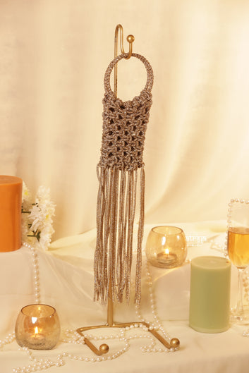 A brown crochet halter bag with long fringe, displayed on a gold stand against a draped cream fabric backdrop, surrounded by candles, pearls, and a glass of champagne.