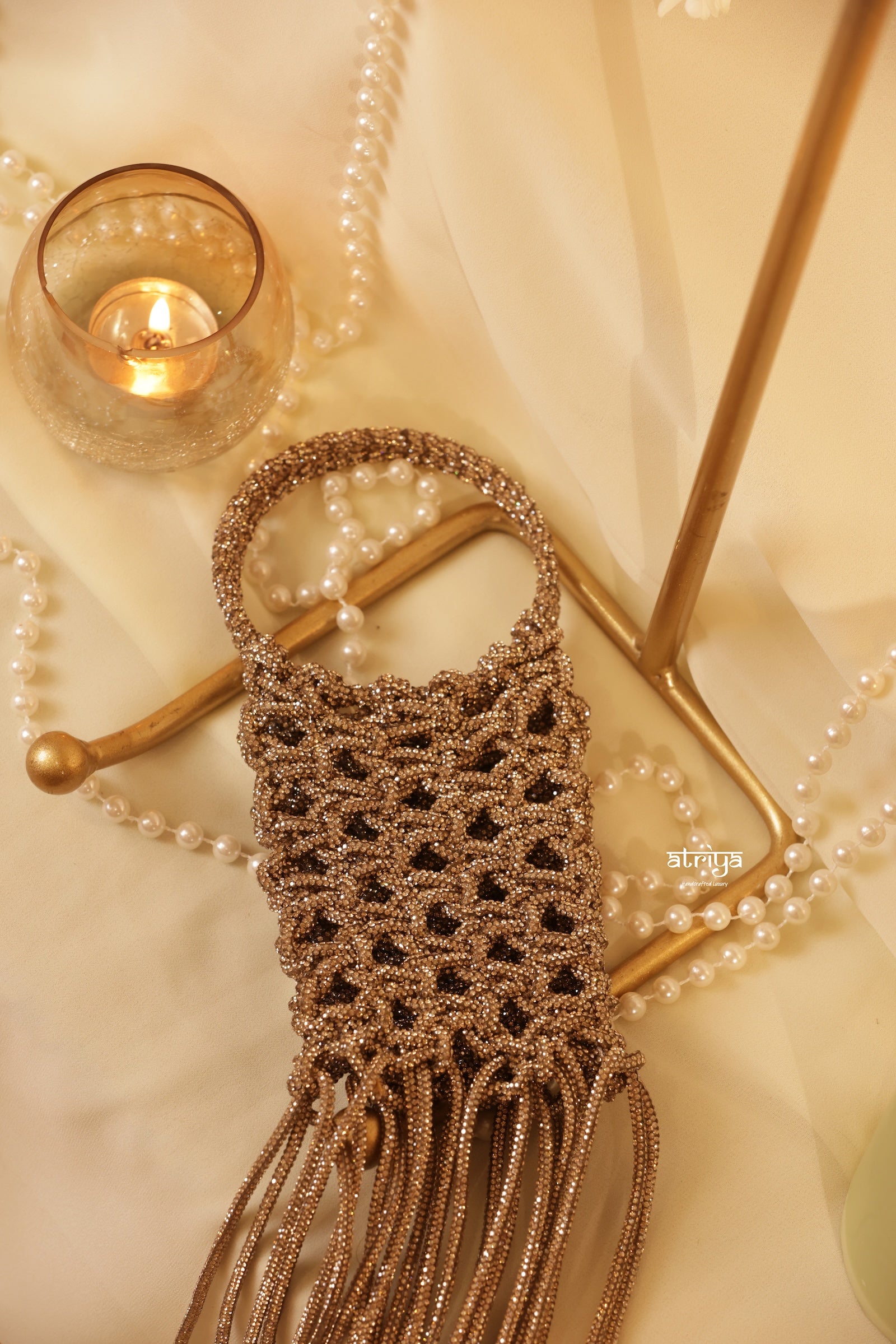 A small woven macrame-style shoulder bag in warm taupe and brown tones, styled on a gold stand with draped cream fabric, pearl strands, and a lit candle in a glass holder.