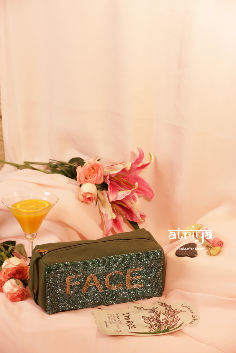 Olive green handcrafted skincare kit with dark glitter beadwork and FACE embroidery, styled with gua sha, jade roller, and pink florals for Atriya Jaipur.
