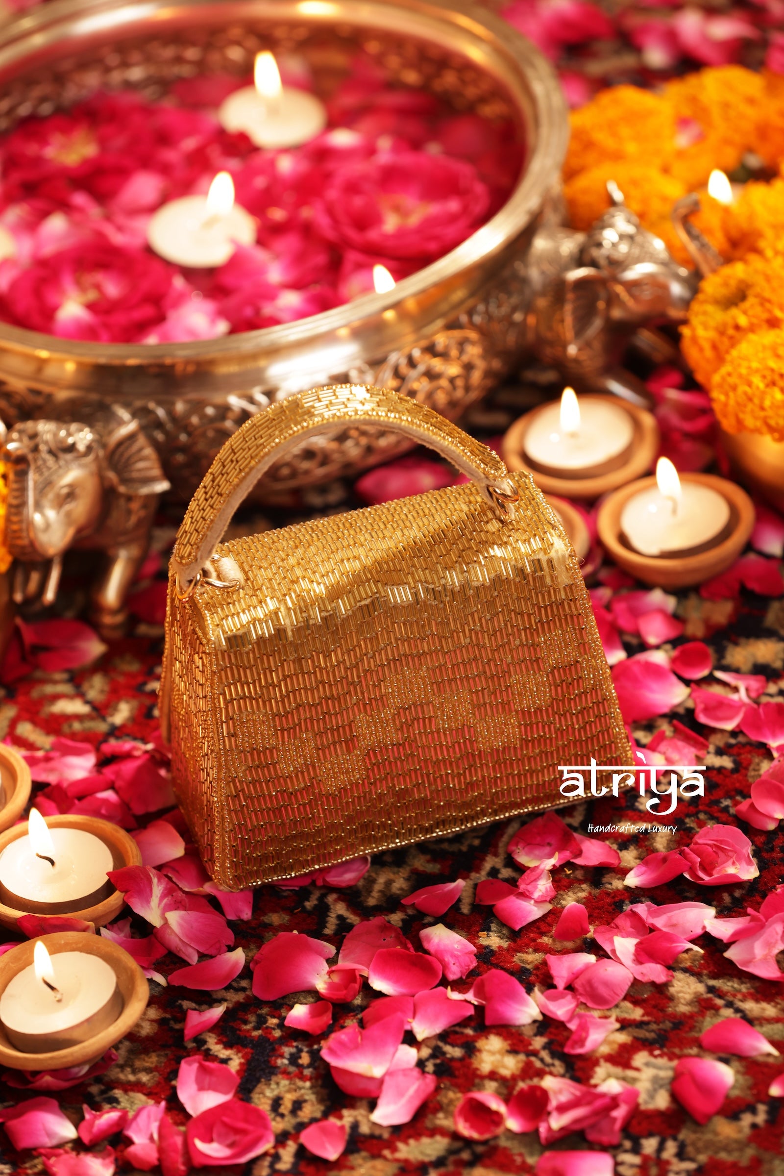 A small metallic gold handbag with a textured woven pattern, styled on a floor covered in pink rose petals with lit candles, a brass bowl of rose petals, and orange marigold flowers. A watermark logo 'atriti' is visible on the bottom right.