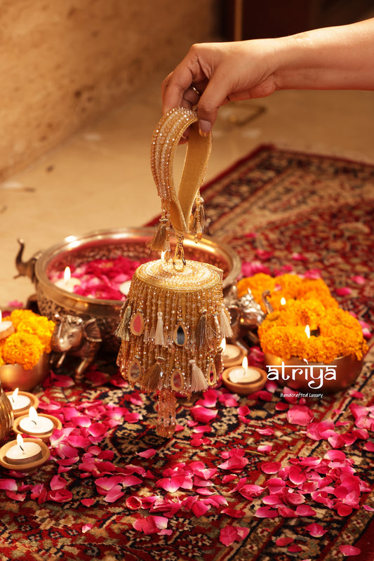 Atriya Noorani Gold handcrafted mirrorwork and tassel fringe cylindrical clutch held by hand strap on floral rug