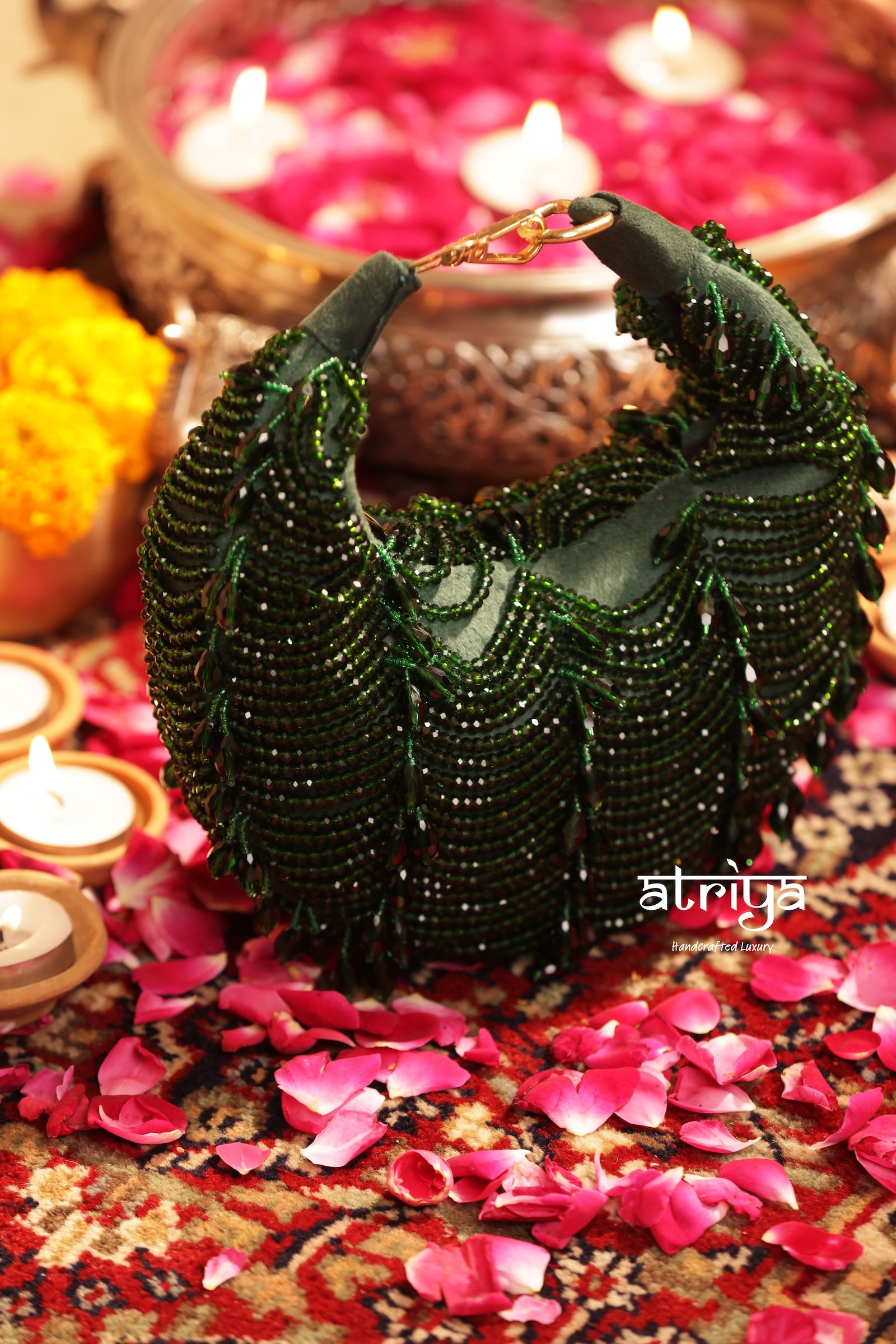 A dark green beaded shoulder bag with a crescent shape, styled on a patterned rug with pink flower petals, lit candles, and marigold flowers in the background.