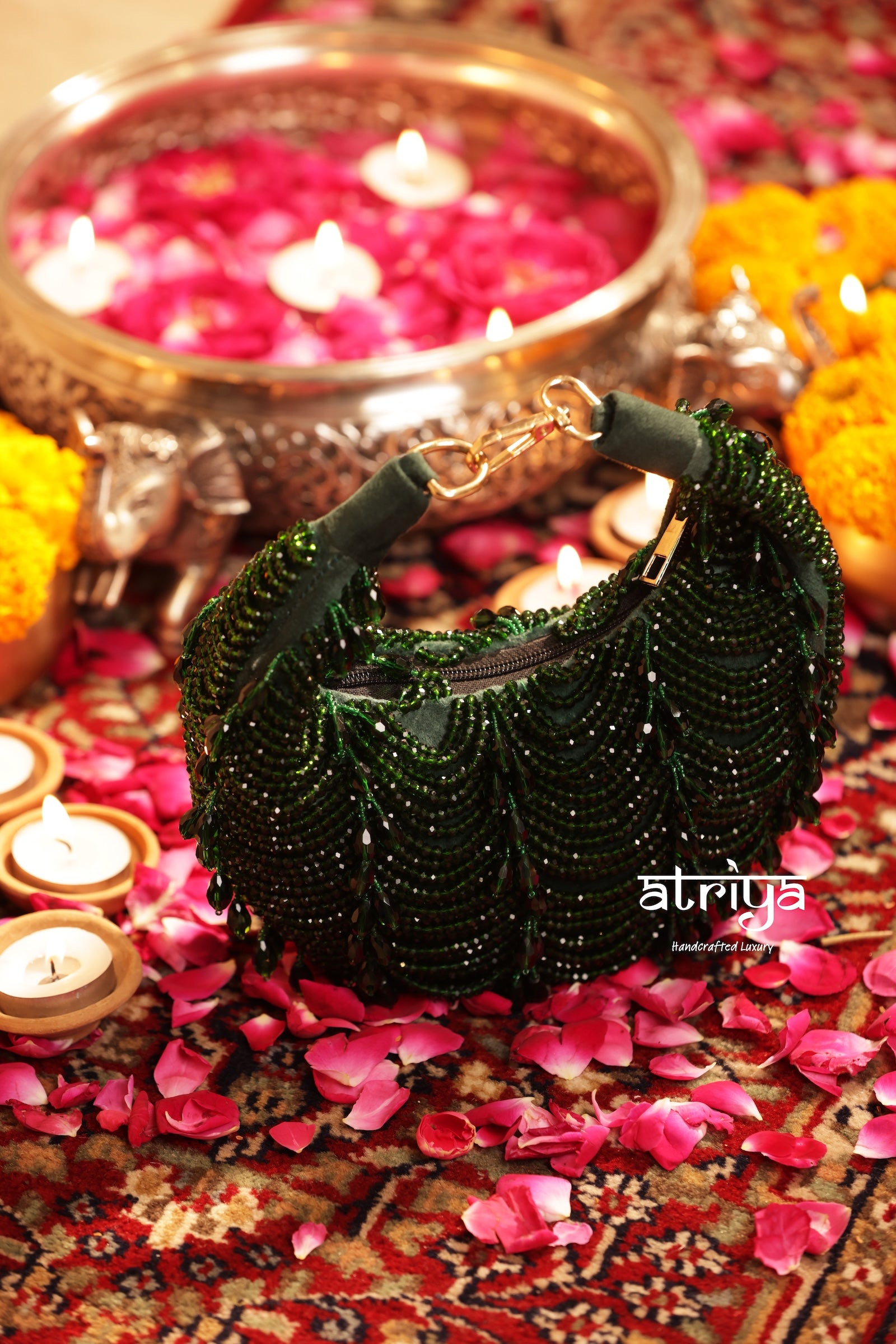A dark green beaded crescent clutch bag with gold-tone hardware, styled on a patterned red carpet surrounded by pink flower petals, lit candles, and a brass oil lamp in the background.
