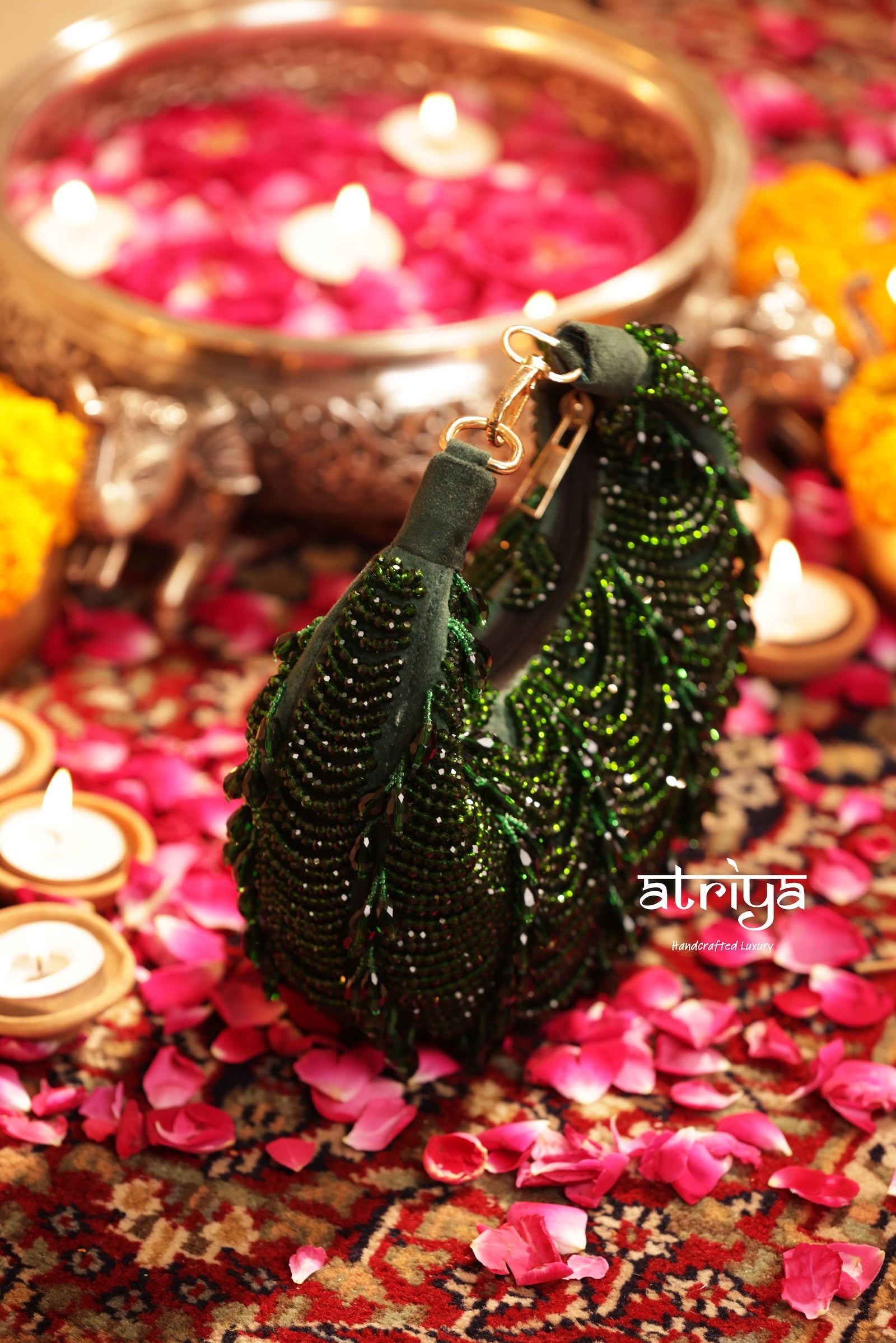 A dark green beaded crescent clutch bag with gold-tone hardware, styled on a patterned rug surrounded by pink flower petals, lit candles, and a brass oil lamp.