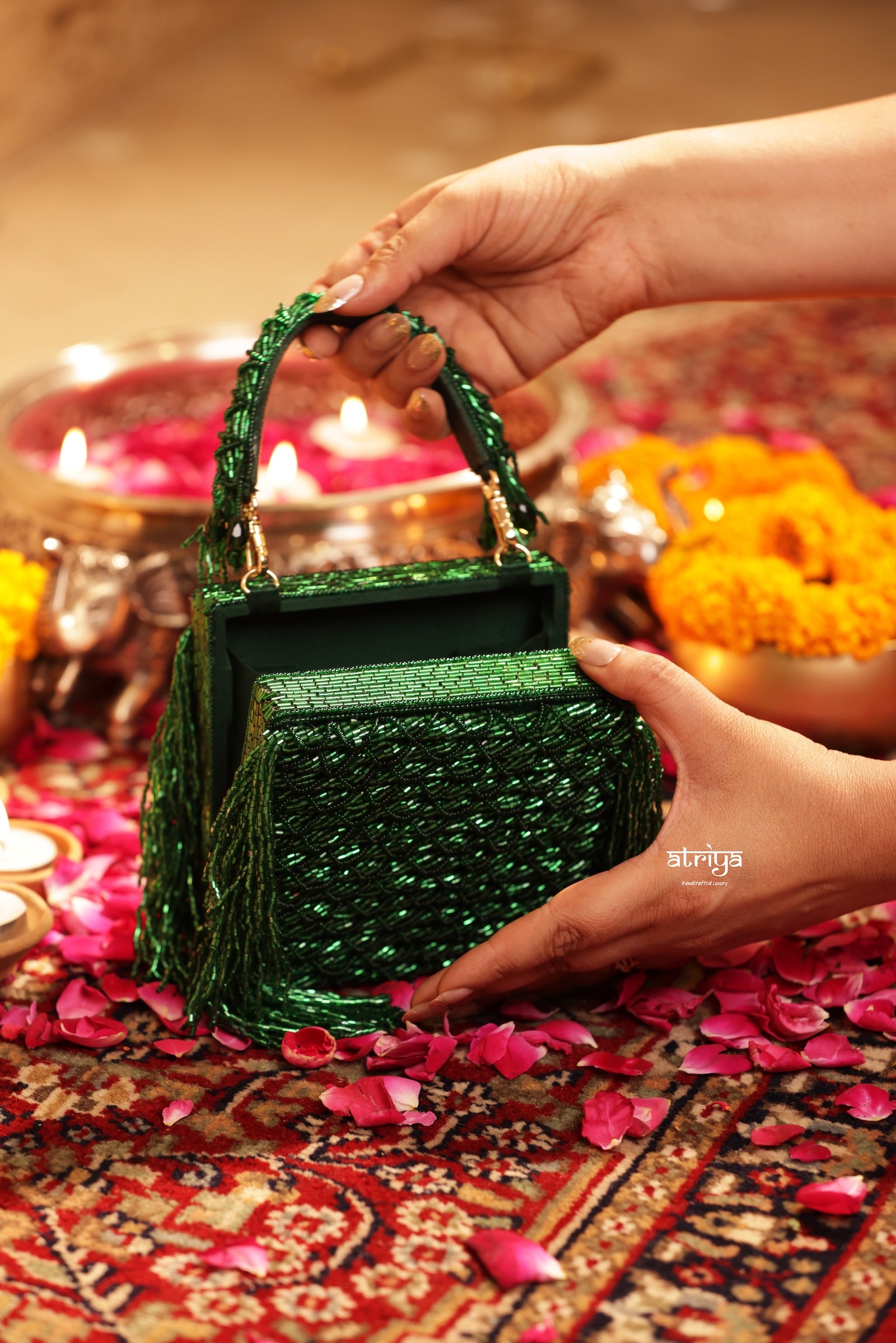 A sparkling green sequined handbag with a top handle and tassel fringe, held by two hands over a decorated festive surface with marigold flowers and lit candles.