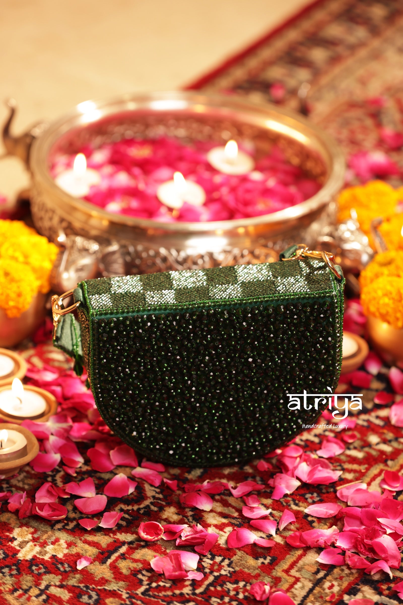 A dark green beaded handbag with a checkered green and white patterned top, styled on a patterned rug with pink flower petals, marigold flowers, and burning candles.