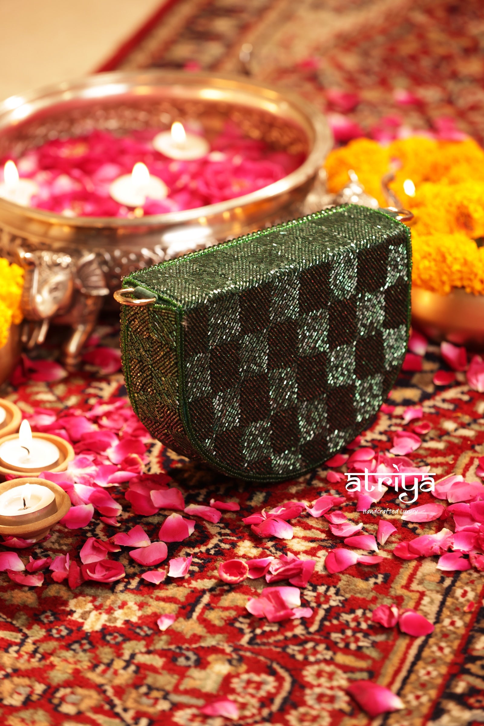 A green and black checkered clutch bag with a metallic sheen, styled on a patterned red rug surrounded by pink flower petals, lit candles, and marigold flowers.