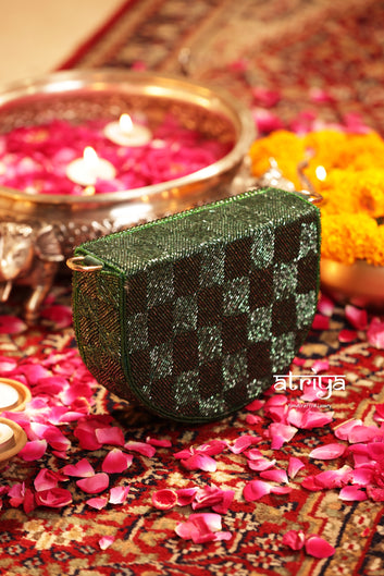 A green and black checkered beaded clutch bag styled on a red patterned rug with pink flower petals, surrounded by lit candles and marigold flowers.