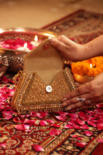 A close-up of a person holding an open gold embellished clutch bag on a patterned rug with pink rose petals, lit candles, and orange marigold flowers in the background.